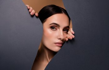 Woman with smooth skin and dark hair parted in the middle peeks through a jagged opening in a dark paper background.